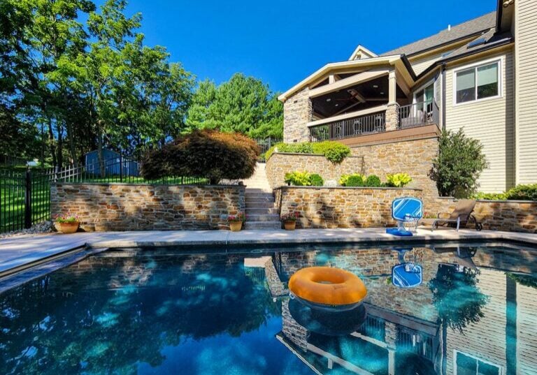 A backyard pool with a yellow inflatable ring, adjacent to a house with a stone exterior, a balcony, stairs, and lush green trees in the background.
