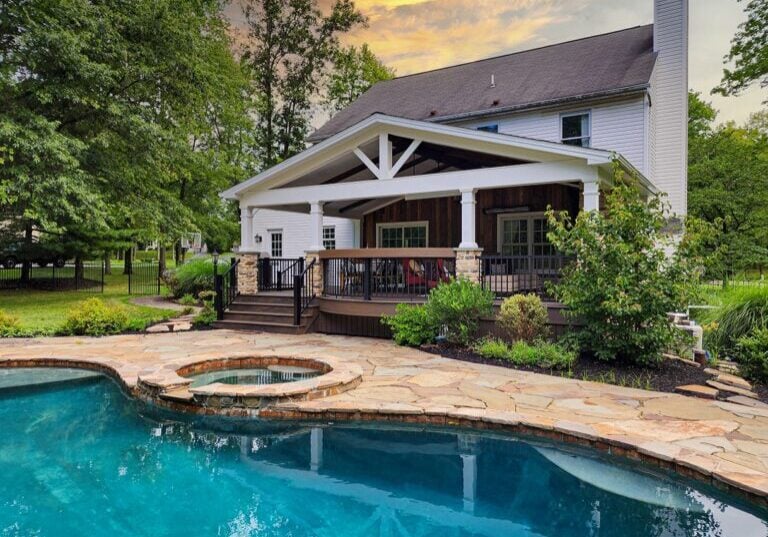 Backyard with a two-story house featuring a covered deck, surrounded by lush greenery and a large swimming pool with a built-in hot tub.