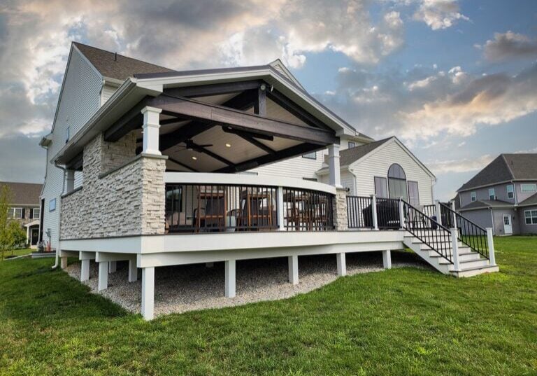 A modern house with an elevated patio featuring stone accents, a slanted roof, and outdoor seating, surrounded by a green lawn under a partly cloudy sky.