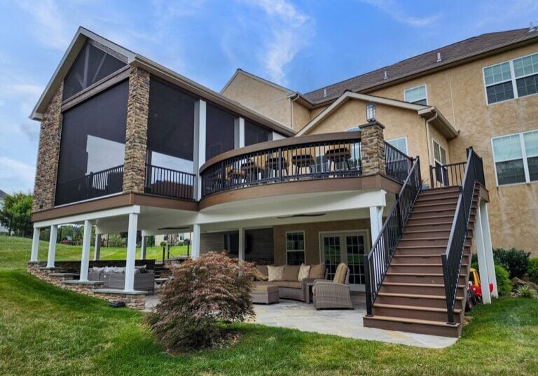 A multilevel house with an outdoor patio, stone columns, an upper deck with railing, screened area, wooden stairs, and lawn furniture on a grassy yard.